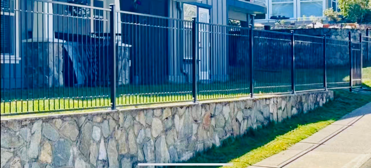 Black metal fence atop a stone retaining wall bordering a residential lawn and sidewalk