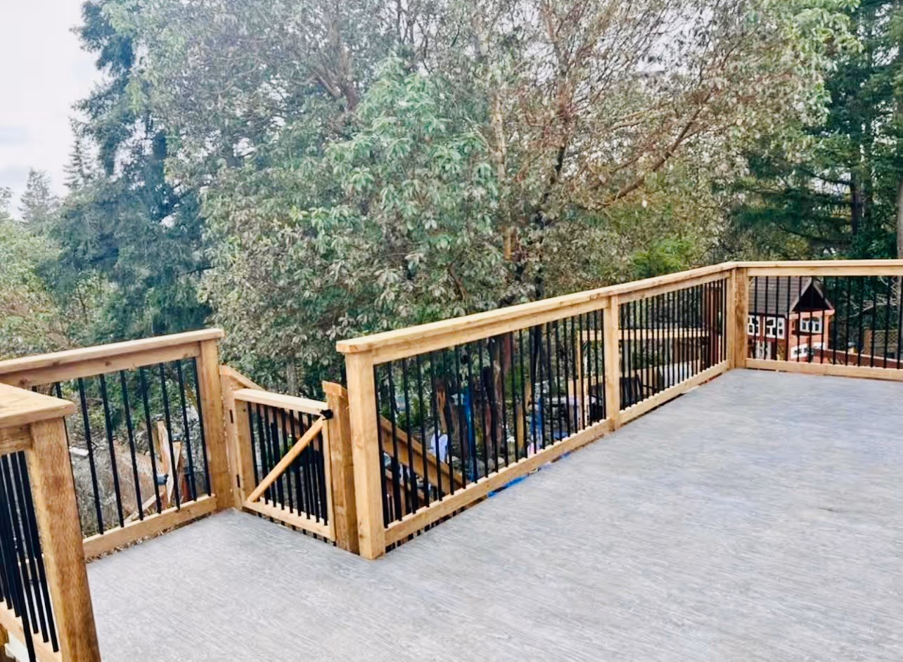 Spacious backyard deck with wood railing, black balusters, and a small gate overlooking trees.